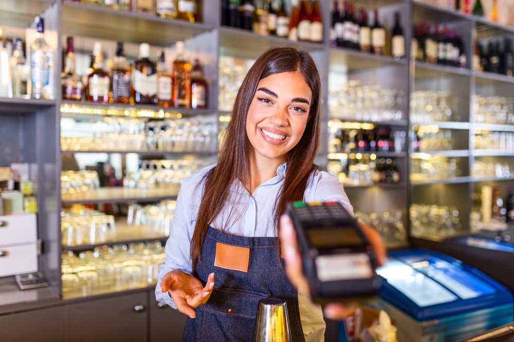 A smiling server in an apron holding out a card terminal across a bar counter